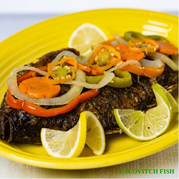Jamaican Escovitch Fish, a whole fried fish topped with pickled vegetables and lemon slices on a yellow plate.